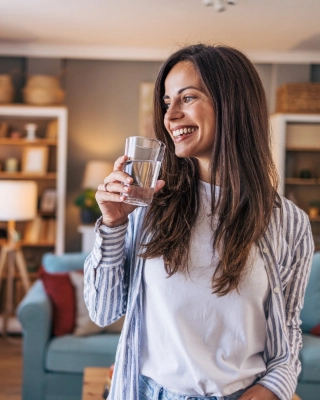 Junge Frau steht im Wohnzimmer und hat Wasserglas in der Hand